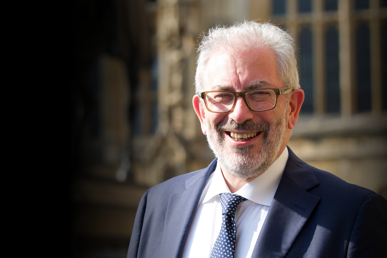Lord Kerslake, a white man in a suit and tie, with glasses and short white hair looks at the camera smiling