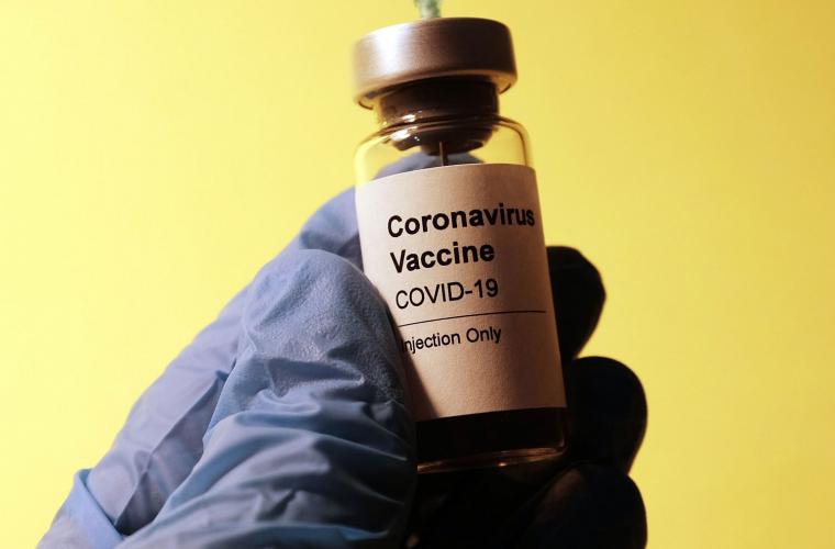 A close up of a gloved hand holding a corona vaccine vial against a yellow background