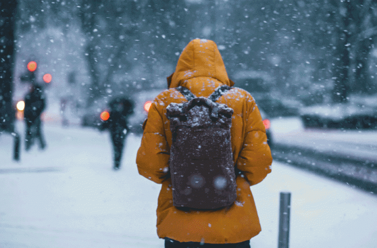 Man with orange jacket and a black backpack walking along a snowy street 