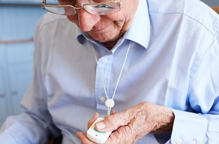 An older man in a shirt and glasses holds a pendant alarm in his hand