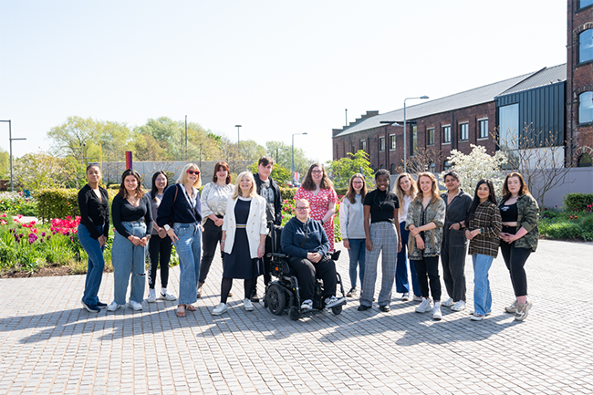 Mayor Tracy Brabin poses with a cohort of 15 Beyond Brontës participants. They are standing on a cobbled courtyard and it is a sunny day.