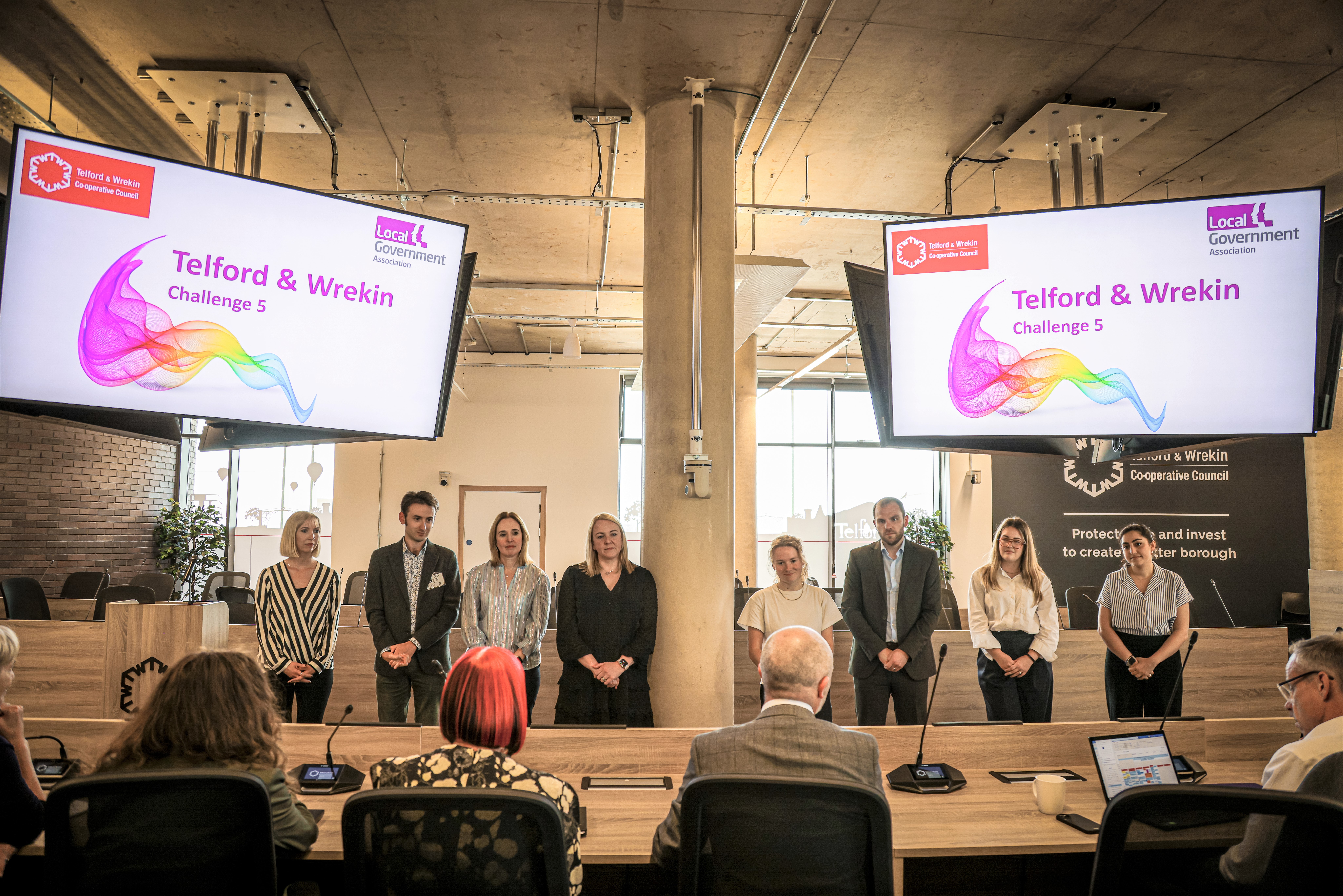 Officers from Telford and Wrekin Council are stood before a seated judging panel. Two large TV screens are above them showing a colourful presentation slide. The text on the slide reads: Telford and Wrekin Challenge 5. There is a rainbow pattern effect below this text as well as the LGA logo and the Telford and Wrekin logo.