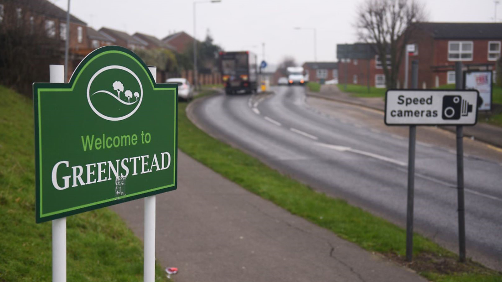 Sign saying "welcome to Greenstead" next to a road leading to a housing estate