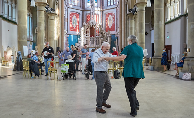 A carer and an elderly gentleman dancing together, behind them is a large circle of musicians, carers and people with dementia dancing and playing instruments in a music group