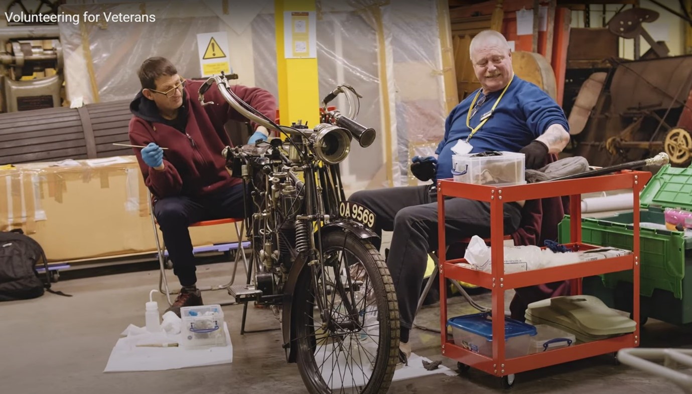 Two white male volunteers sit either side of a vintage motorbike from Birmingham Museum Trust’s collection, they are both in their late 50’s. 