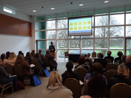 View from the back of a hall of people in a meeting looking at a presentation