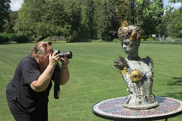 – A woman in black clothes stands on the left holding a camera to her eye. She is photographing a sculpture, which is on the right of the image. 