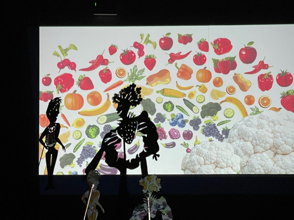 A shadow puppet performance with a adult and child puppet with various fruits and vegetables in the background.