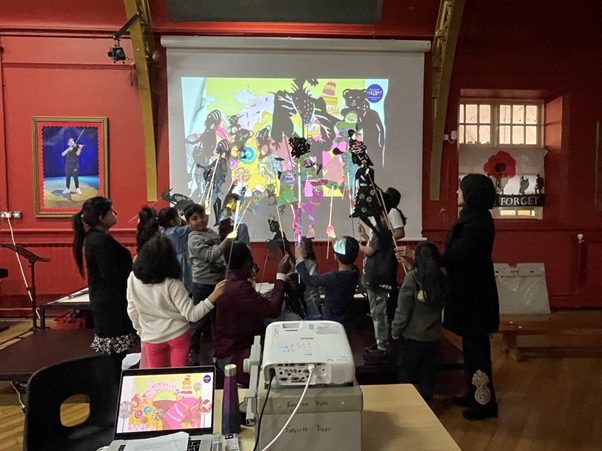 A group of children with a teacher gathered around performing a shadow puppet performance in front of a projected screen with animals and people puppets.