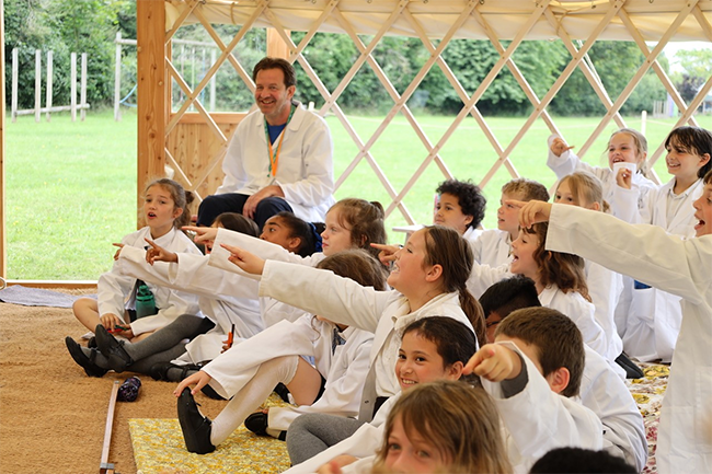 A class of children are sat in a Mongolian yurt wearing white lab coats looking towards the stage.