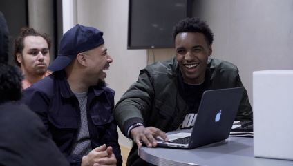 Two men are looking towards each other and laughing. They are sat behind a laptop and one of the men is pressing buttons on the laptop. 