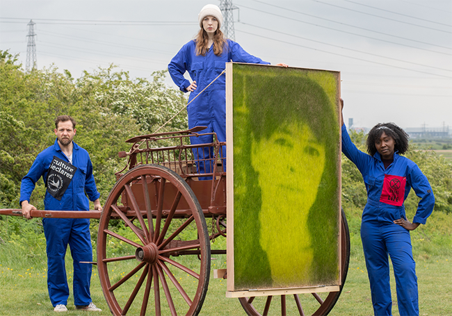 There is an old-fashioned cart in a field, with a large photograph of a woman. Three people wearing blue boiler suits are standing around the cart. 