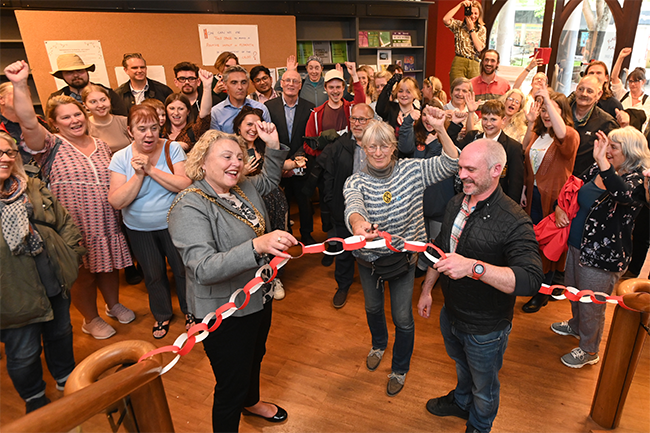 A large crowd of people are cheering with their hands in the air as they watch two women and a man cut a ribbon to officially open the Climate Hub in a disused shop in Plymouth City Centre.    