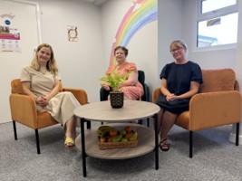 A room with light grey paint and a rainbow decal on the walls. Sat around a coffee table are three women, each on a comfy chair. From left to right): Louise Moriarty - Family Hub Practitioner, Lee Stacey - Business Support Officer, Ileana Monaghan - Family Hub Navigator