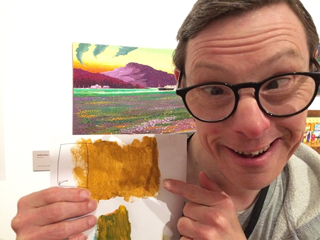 Richard, an artist from The Professors, holds up a piece of paper showing two mustard-coloured painted rectangles. 