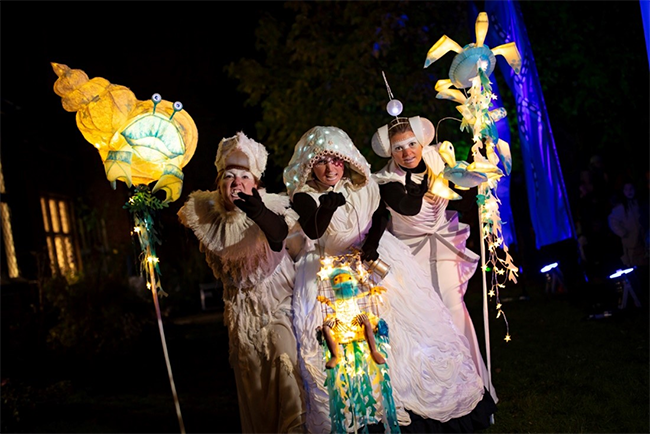 Three performers wearing Pilgrim costumes and model boats with sails and lights look out to the sky holding telescopes in the dark outside