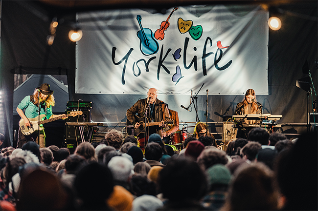 A festival stage with large backdrop, reading York Life, in front of a big crowd. Four members of the band Bull are performing with a variety of musical instruments.