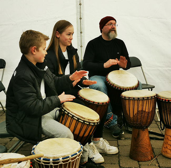 Three people, two young and one older, take part in a drumming workshop inside a marquee with bunting.  