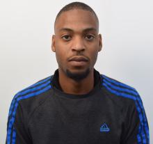 Ackeem is stood against a white background wearing a black Adidas tracksuit with blue stripes