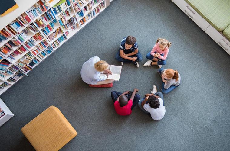 Children and a female adult sitting in a circle inside a library. The adult is reading to the children.