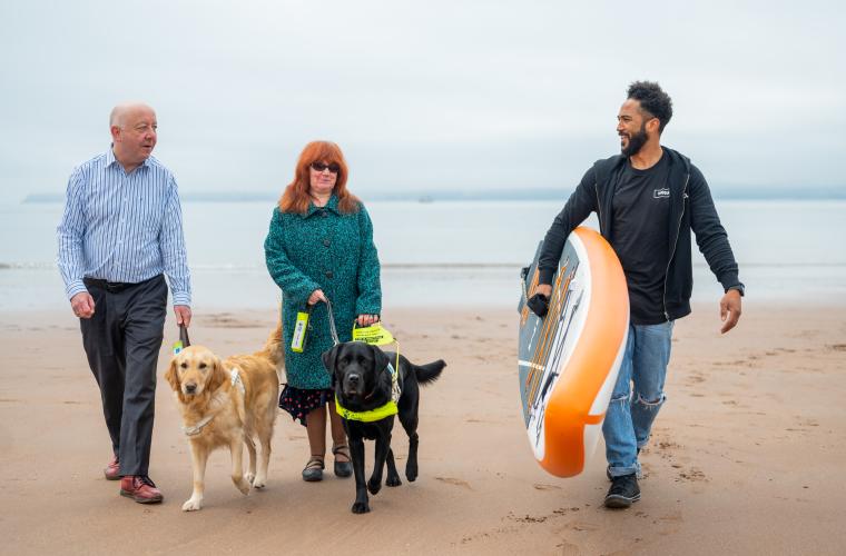Steve and Mandy walking with their guide dogs on the beach alongside a man with a surfboard
