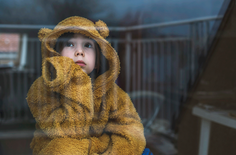 A child dressed in a bear jumpsuit staring sadly at the window.