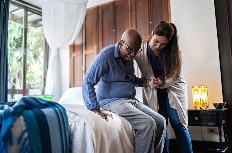 An adult woman helping an elderly to stand from his bed