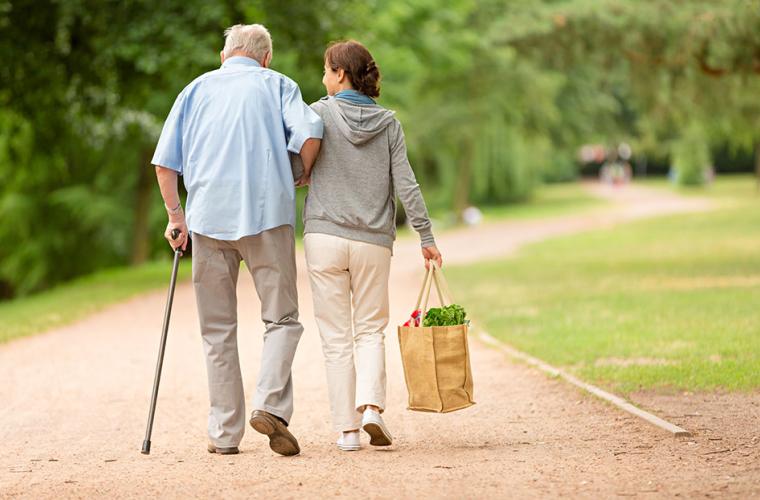 An elderly and a carer walking side by side. The elderly is using a crutch and holding the carer arm, while she carries some food as well.