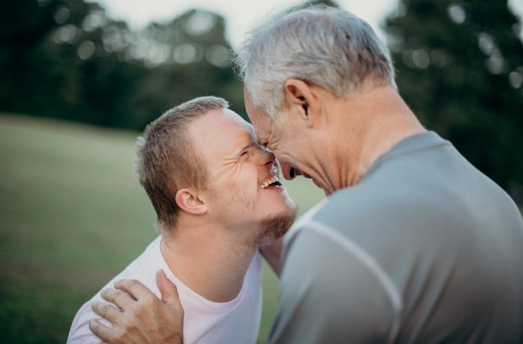 Two men, one older and one younger with Down's Syndrome, smiling at each other on a grass field
