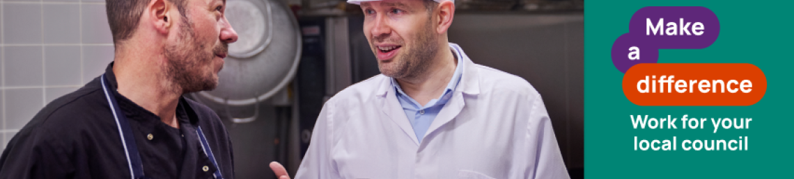 two men in a industrial kitchen. One is wearing an apron and the other is white, looking like a supervisor