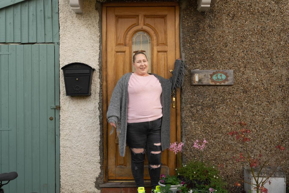 A white lady with a pink top and grey cardigan stands in front of a wooden house door and a brick wall. The other side is a black metal post box.