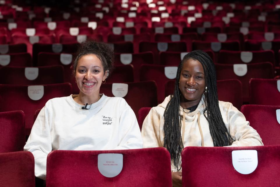 two black women sit in red velvet theatre chairs facing the camera smiling. Both are wearing light sweatshirts.