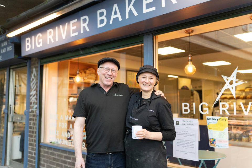 A white couple stand in front of a bakery with their arms round each other smiling