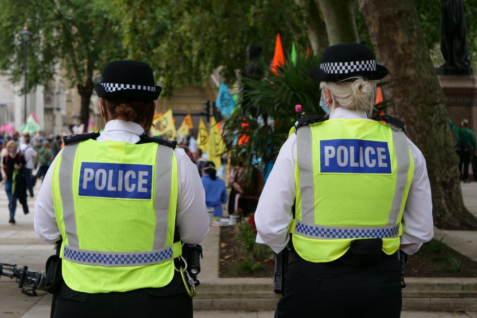 Two woman police officers with their back to the camera, they have hi-vis on which says Police on them and in the background they are watching a protest in a square 